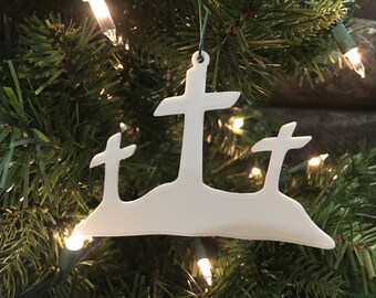 Könnte beinhalten: Weißer Weihnachtsschmuck mit drei Kreuzen auf einem Hügel. Der Schmuck besteht aus einem einfarbigen weißen Material und wird an einer grünen Schnur aufgehängt. Der Hintergrund ist ein Weihnachtsbaum mit Lichtern.