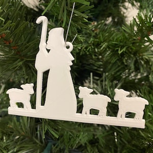 Puede incluir: Adorno navideño de silueta blanca de un pastor con un bastón y cuatro ovejas. El adorno está colgado de un árbol de Navidad verde.