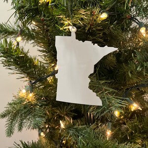 Può includere: Addobbo bianco a forma dello stato del Minnesota appeso a un albero di Natale verde con luci bianche.