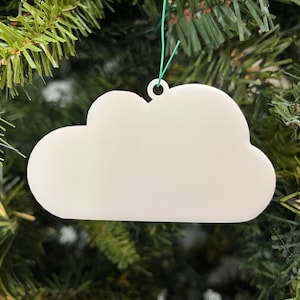 Puede incluir: Adorno de Navidad blanco en forma de nube con un agujero para colgar. El adorno está hecho de un material blanco liso y está colgado de un árbol de Navidad verde.