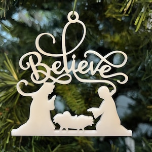 May include: White Christmas bauble featuring a nativity scene and the word "Believe" in a decorative script.