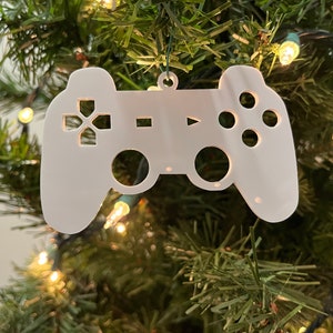 Puede incluir: Adorno navideño de plástico blanco con forma de mando de videojuego. El adorno tiene un diseño de mando tradicional con botones y una cruz direccional.