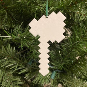 May include: White pixelated pickaxe Christmas ornament hanging on a green artificial Christmas tree.