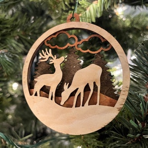May include: A wooden Christmas ornament featuring a laser-cut design of two deer standing in a snowy forest scene with trees and clouds. The ornament is made of light brown wood and has a rustic, natural look.