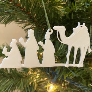 May include: White 3D printed Christmas ornament depicting the three wise men and a camel. The ornament is hanging on a Christmas tree.