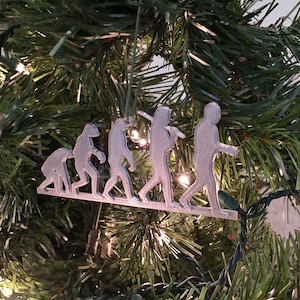 May include: A silver-colored Christmas ornament depicting the stages of human evolution. The ornament features a series of figures, starting with a primitive ape-like creature and progressing to a modern human.
