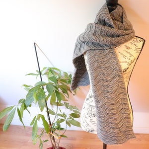 Puede incluir: Una bufanda de ganchillo gris con un patrón ondulado, expuesta en un maniquí blanco.
