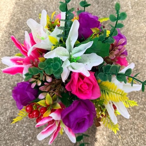 May include: A floral arrangement featuring white lilies, vibrant hot pink roses, and deep purple roses. The bouquet includes green eucalyptus-style foliage, yellow fern-like leaves, and small red berries, creating a dense, multi-coloured botanical display.