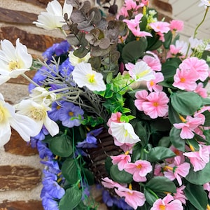 May include: A hanging basket filled with artificial flowers in shades of white, pink, and purple, with green leaves. The basket is made of woven brown material and is suspended from a hook. The flowers create a vibrant display.