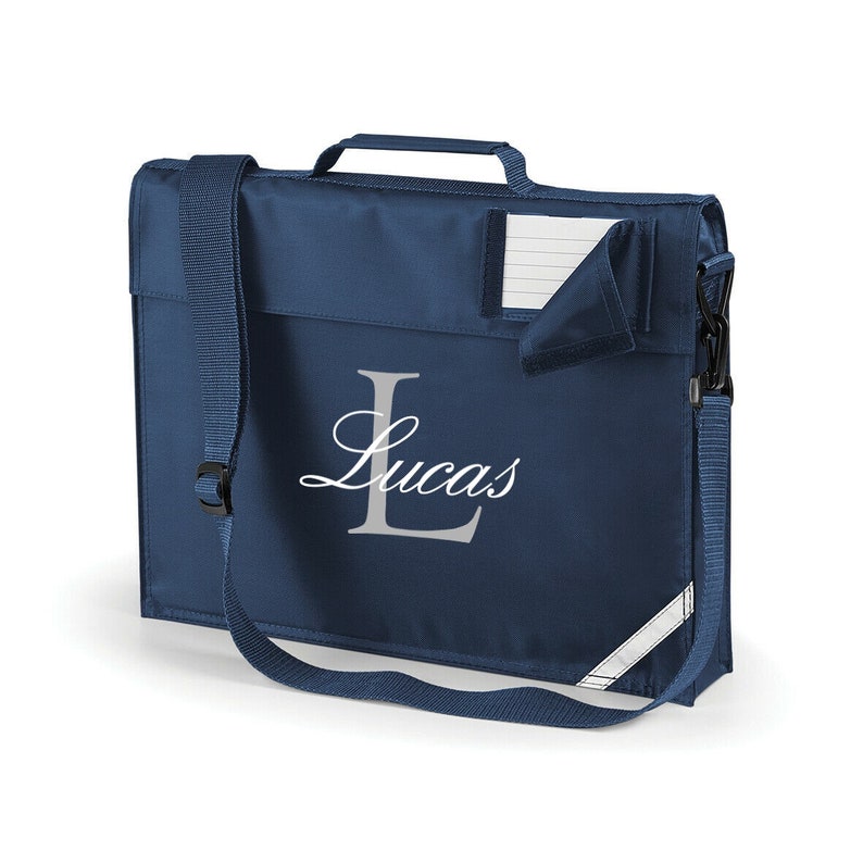 Pu&ograve; includere: Uno zaino blu navy con una tracolla regolabile bianca e una striscia riflettente bianca. Lo zaino ha un manico bianco e una tasca bianca sul davanti con un blocco note bianco a righe all'interno. Lo zaino &egrave; personalizzato con una "L" bianca in corsivo e il nome "Lucas".