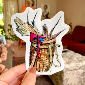 May include: A hand-drawn illustration of a wicker basket with a red and blue axe, antlers, and sticks inside. The basket has a strap and is drawn in a rustic style.
