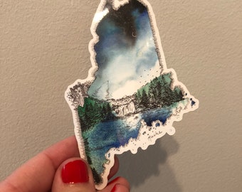 May include: A sticker of the state of Maine with a watercolor-style illustration of a forest, lake, and mountains. The sticker has a blue, green, and white color scheme and the word "Maine" is written in the bottom right corner.