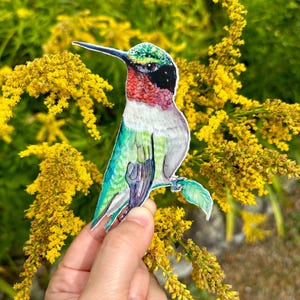 May include: A paper cutout of a hummingbird with green, blue, red, and black feathers. The hummingbird is perched on a branch with green leaves. The cutout is held in front of a background of yellow flowers.