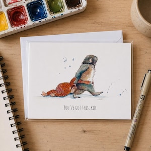 May include: A white greeting card featuring a watercolour illustration of a large grey creature walking while carrying a smaller orange creature on its back. The text "YOU'VE GOT THIS, KID" is printed in black below the illustration on the card.