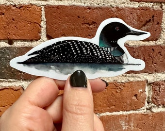 Könnte beinhalten: Ein schwarzer und weißer Loon-Aufkleber mit blauem und grünem Kopf. Der Aufkleber befindet sich auf einem Ziegelhintergrund.