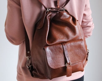 May include: A brown leather backpack with a flap closure and a buckle. The backpack has a large main compartment and a smaller front pocket. The backpack is being worn by a person wearing a pink hoodie.