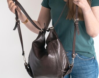 May include: A dark brown leather backpack with a slouchy, unstructured silhouette. The bag features thin, adjustable shoulder straps with silver-toned metal buckles and a vertical zippered pocket on the front exterior. The material has a smooth, slightly glossy finish.