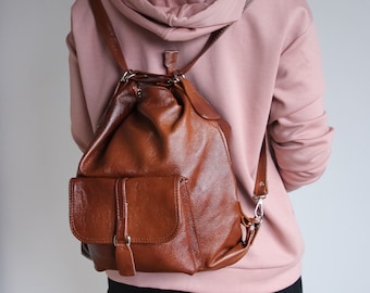 May include: A brown leather backpack with a textured surface, featuring a drawstring closure, a front flap pocket with a vertical strap detail, and adjustable shoulder straps. The bag has a soft, slouchy structure and silver-toned metal hardware.
