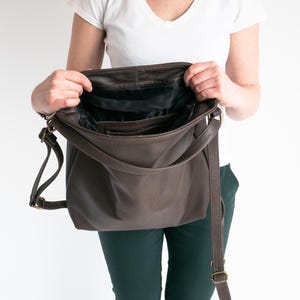 May include: A dark brown leather shoulder bag with an open top, revealing a black fabric interior lining. The bag features a long, adjustable shoulder strap with antique brass-toned metal hardware and a zippered pocket inside the main compartment.