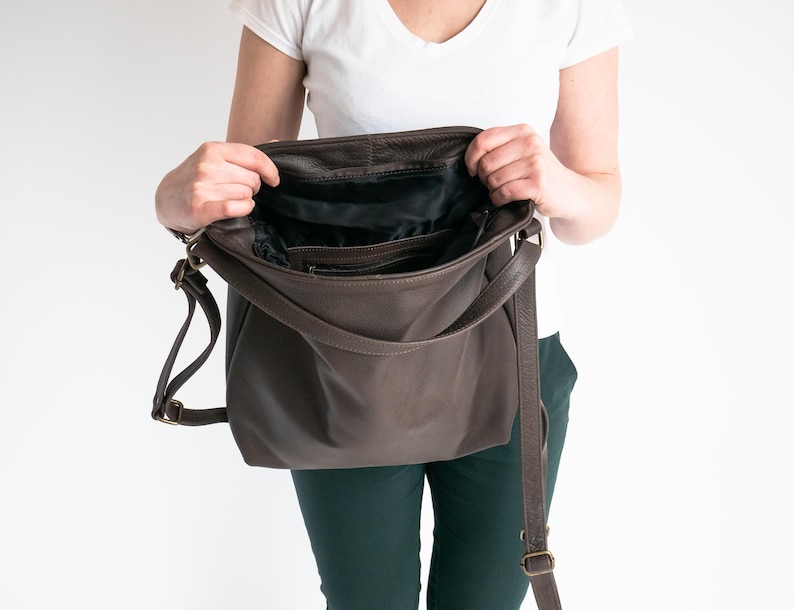 May include: A dark brown leather shoulder bag with an open top, revealing a black fabric interior lining. The bag features a long, adjustable shoulder strap with antique brass-toned metal hardware and a zippered pocket inside the main compartment.