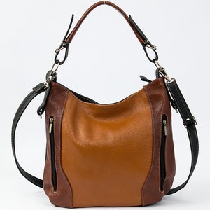 Puede incluir: Un bolso de mano de cuero marr&oacute;n con una correa negra y un bolsillo con cremallera. El bolso tiene una combinaci&oacute;n de colores marr&oacute;n, negro y beige.