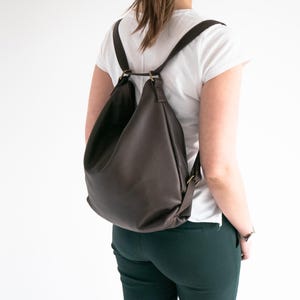 May include: A dark brown leather backpack with a slouchy, unstructured silhouette. The bag features thin shoulder straps attached to brass-toned metal rings at the top, creating a gathered opening. The material has a visible pebbled texture.
