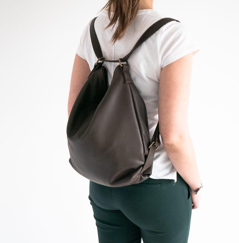 May include: A dark brown leather backpack with a slouchy, unstructured silhouette. The bag features thin shoulder straps attached to brass-toned metal rings at the top, creating a gathered opening. The material has a visible pebbled texture.