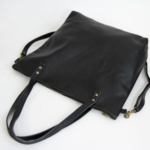 May include: A black tote bag with a textured surface, featuring two top handles secured by brass-toned rivets. The bag includes a top zipper closure and a detachable shoulder strap with brass-toned swivel hooks on each side.