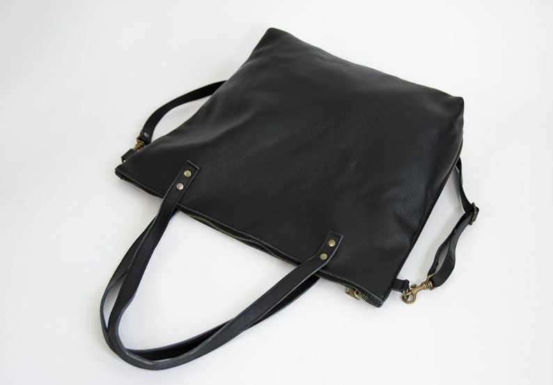 May include: A black tote bag with a textured surface, featuring two top handles secured by brass-toned rivets. The bag includes a top zipper closure and a detachable shoulder strap with brass-toned swivel hooks on each side.