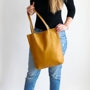 May include: A mustard yellow leather tote bag with white leather trim and handles. The bag is being held by a person wearing blue jeans.