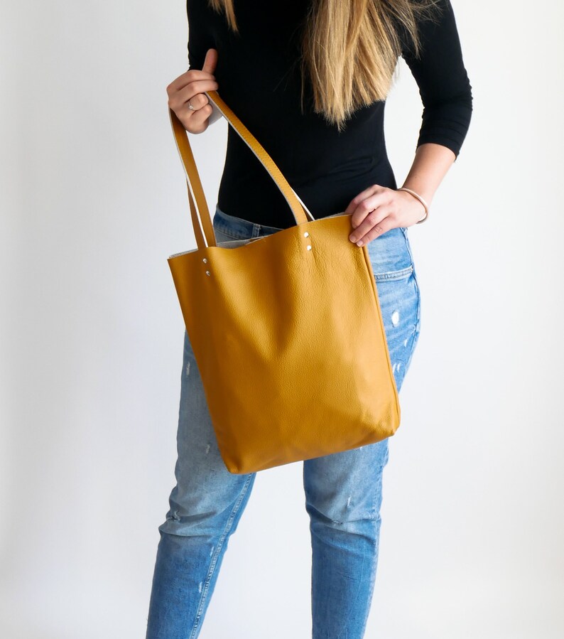 May include: A mustard yellow leather tote bag with white leather trim and handles. The bag is being held by a person wearing blue jeans.