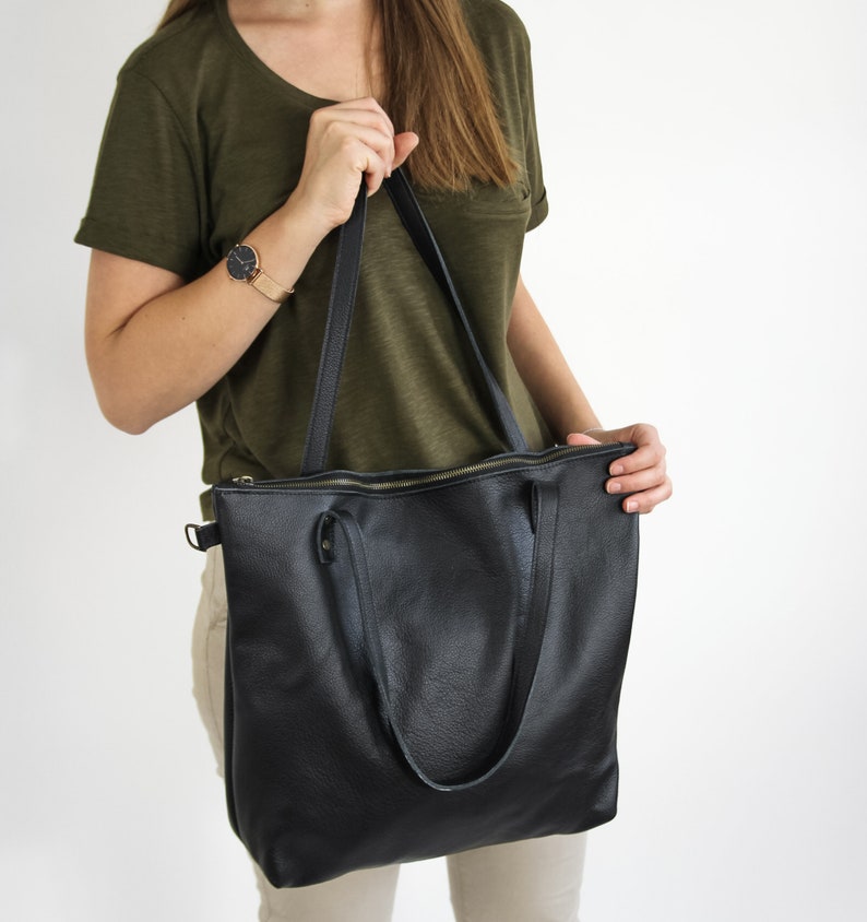 May include: A black leather tote bag with a textured surface, featuring two long shoulder straps, a top zipper closure with a metallic pull, and a small side D-ring attachment for additional utility or strap options.