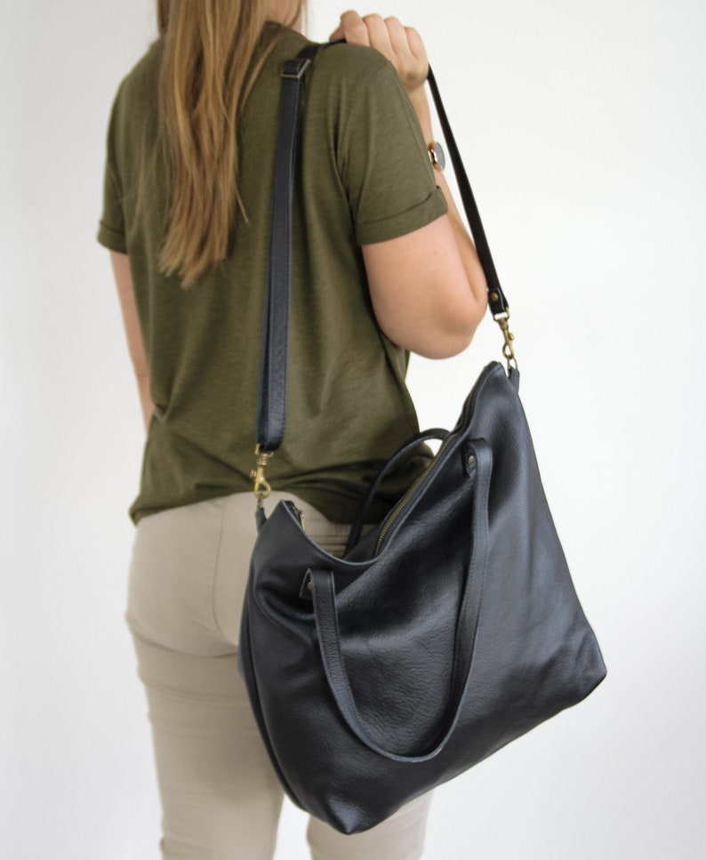 May include: A black leather tote bag with a textured surface, featuring two top handles and a detachable, adjustable shoulder strap with brass-toned metal hardware. The bag has a zippered top closure and a spacious, slouchy silhouette.
