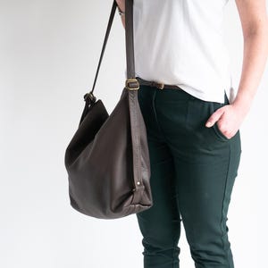 May include: A dark brown leather hobo bag with a slouchy silhouette, featuring a long adjustable shoulder strap with antique brass-toned metal hardware buckles and a smooth, pebbled texture throughout the exterior surface.