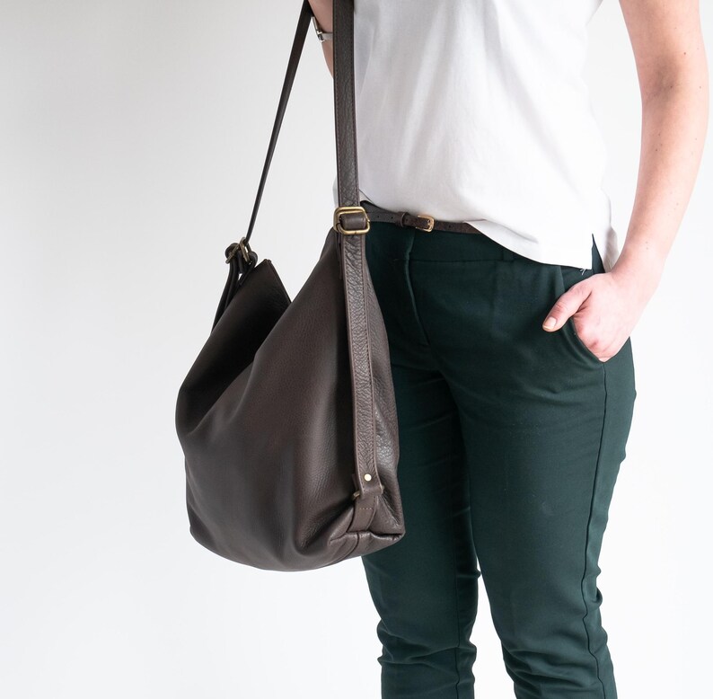 May include: A dark brown leather hobo bag with a slouchy silhouette, featuring a long adjustable shoulder strap with antique brass-toned metal hardware buckles and a smooth, pebbled texture throughout the exterior surface.