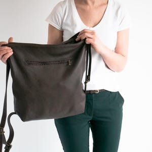 May include: A dark brown leather hobo bag featuring a horizontal zippered pocket on the front exterior, a main top zipper closure, and adjustable shoulder straps with antique brass-toned metal hardware buckles and sliders.