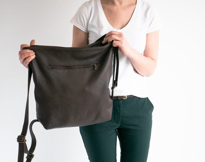 May include: A dark brown leather hobo bag featuring a horizontal zippered pocket on the front exterior, a main top zipper closure, and adjustable shoulder straps with antique brass-toned metal hardware buckles and sliders.