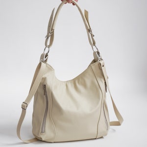 Peut inclure: Un sac &agrave; main en cuir de couleur cr&egrave;me avec une longue bandouli&egrave;re et deux poches zipp&eacute;es &agrave; l'avant.