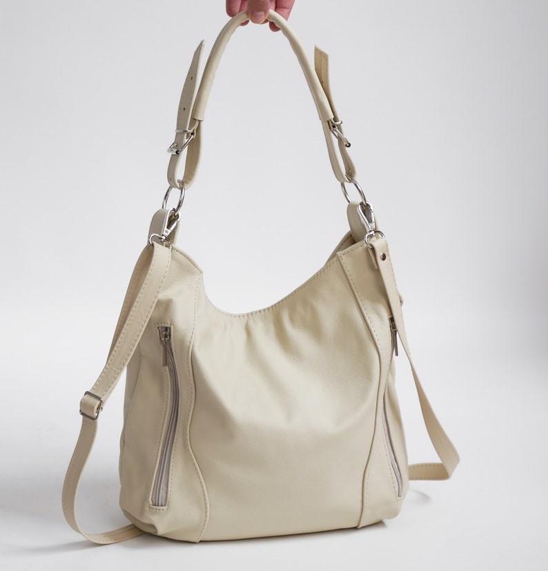 Peut inclure: Un sac &agrave; main en cuir de couleur cr&egrave;me avec une longue bandouli&egrave;re et deux poches zipp&eacute;es &agrave; l'avant.