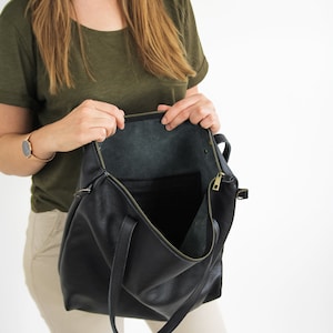 May include: A black leather tote bag with a textured surface, featuring a top zipper closure with a gold-toned metal pull. The interior is open and unlined, showing the natural dark underside of the leather material.