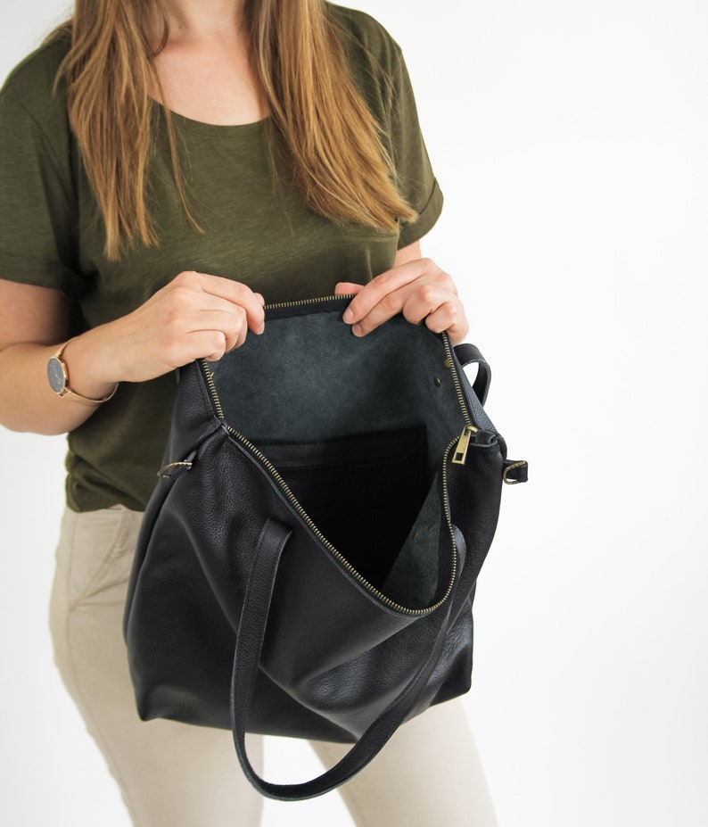 May include: A black leather tote bag with a textured surface, featuring a top zipper closure with a gold-toned metal pull. The interior is open and unlined, showing the natural dark underside of the leather material.