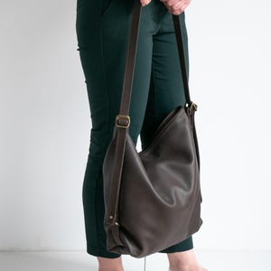 May include: A dark brown leather hobo bag with a slouchy silhouette, featuring adjustable brown straps and antique brass-toned metal hardware. The bag has a smooth, pebbled texture and is worn over the shoulder with a long, thin strap.