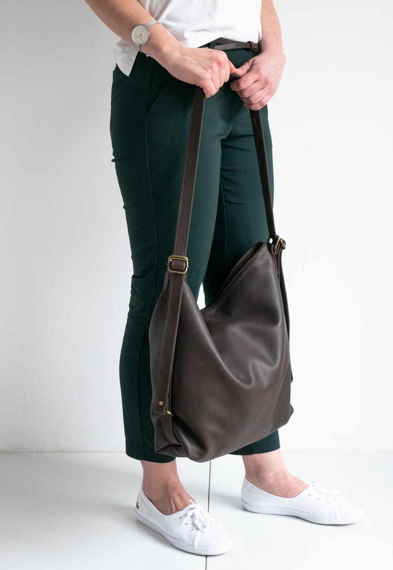 May include: A dark brown leather hobo bag with a slouchy silhouette, featuring adjustable brown straps and antique brass-toned metal hardware. The bag has a smooth, pebbled texture and is worn over the shoulder with a long, thin strap.