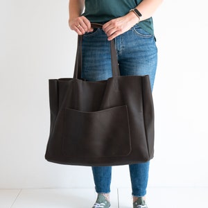 May include: A large, dark brown leather tote bag with a front pocket and long handles. The bag is held by a person wearing blue jeans and green sneakers. The bag appears to be empty.