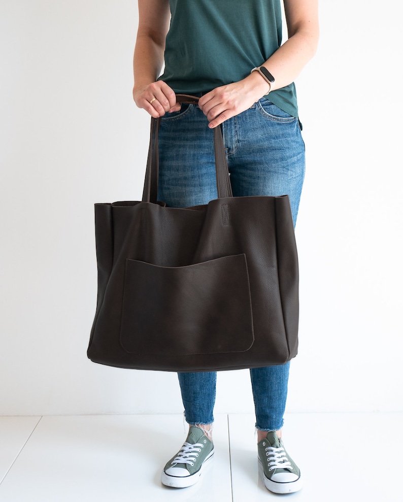 May include: A large, dark brown leather tote bag with a front pocket and long handles. The bag is held by a person wearing blue jeans and green sneakers. The bag appears to be empty.
