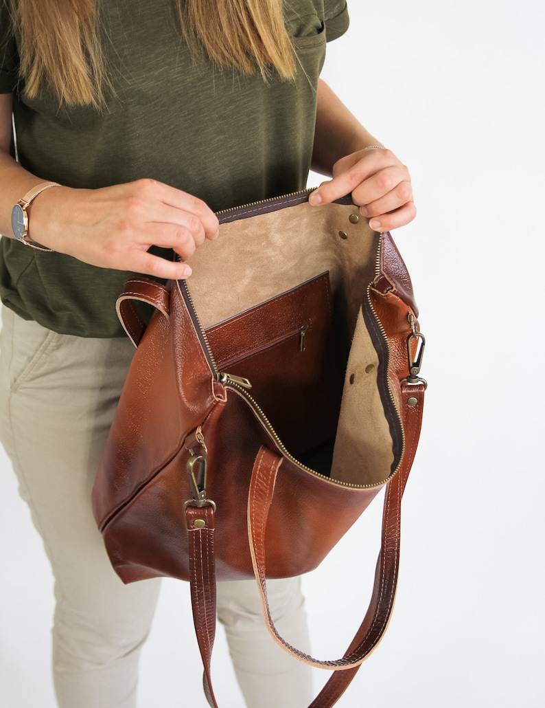 May include: A brown leather tote bag with a textured surface, featuring a top zipper closure, two leather shoulder straps with brass-toned metal clips, and an interior pocket with a zipper. The interior is lined with a light tan material.
