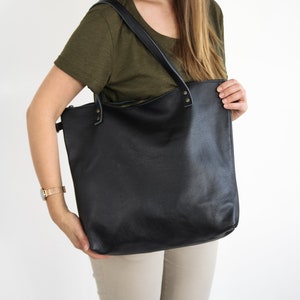 May include: A large black leather tote bag featuring a top zipper closure, two shoulder straps attached with metallic rivets, and a textured surface. The bag has a rectangular shape with rounded bottom corners and a spacious main compartment.