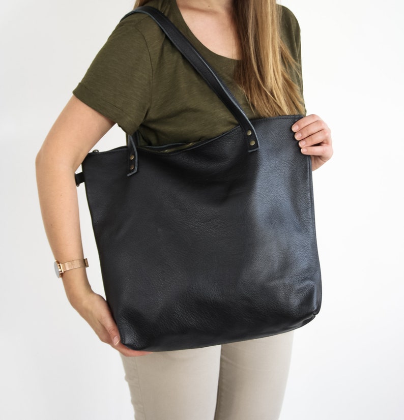 May include: A large black leather tote bag featuring a top zipper closure, two shoulder straps attached with metallic rivets, and a textured surface. The bag has a rectangular shape with rounded bottom corners and a spacious main compartment.