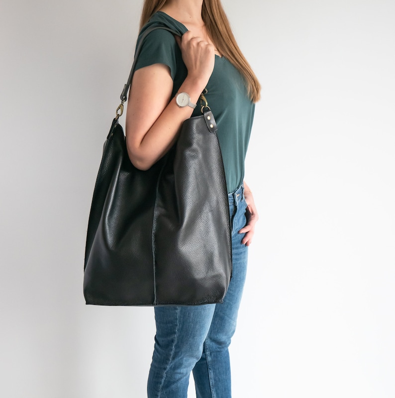 K&ouml;nnte beinhalten: Eine schwarze Ledertasche mit einem langen Trageriemen. Die Tasche wird &uuml;ber der Schulter einer Person getragen, die ein t&uuml;rkisfarbenes Shirt und eine blaue Jeans tr&auml;gt.