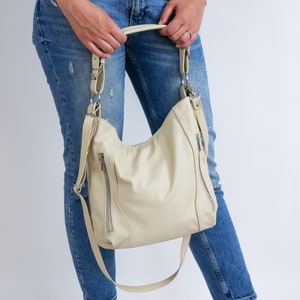 Peut inclure: Un sac &agrave; main en cuir de couleur cr&egrave;me avec des ferrures argent&eacute;es et une fermeture &agrave; glissi&egrave;re. Le sac est port&eacute; par une personne v&ecirc;tue d'un jean bleu.